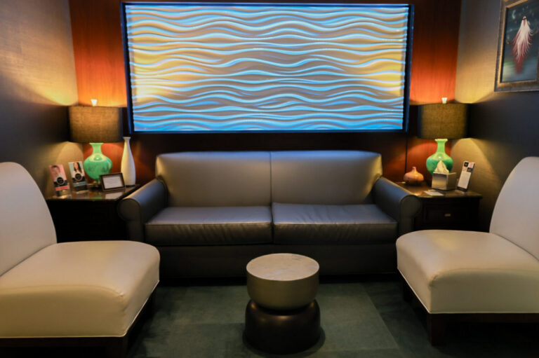 A modern seating area featuring a gray leather sofa flanked by two white armchairs. In the background, a blue and beige wave-patterned wall panel is illuminated. Two green lamps are on side tables beside the sofa, and a small round table sits in the center.